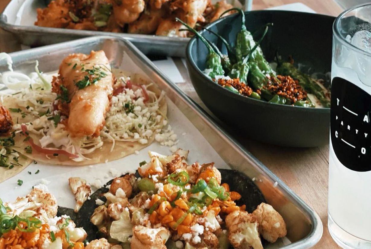 A close up of a plate with fish tacos and crispy cauliflower, next to a bowl of blistered shishito peppers.